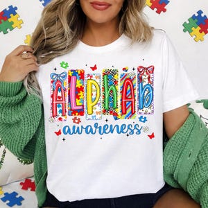 May include: White t-shirt featuring the word "ALPHA" in colourful, puzzle-piece-themed letters, and the word "awareness" in blue script. The shirt design includes butterflies and flowers.