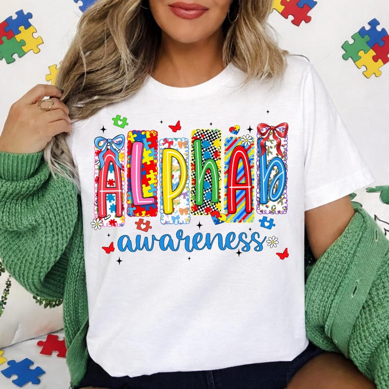 May include: White t-shirt featuring the word "ALPHA" in colourful, puzzle-piece-themed letters, and the word "awareness" in blue script. The shirt design includes butterflies and flowers.