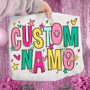 May include: White tote bag with the words "CUSTOM NAME" in colorful, glittery letters. The design includes stars, hearts, and smiley faces. The bag is held up by a person.