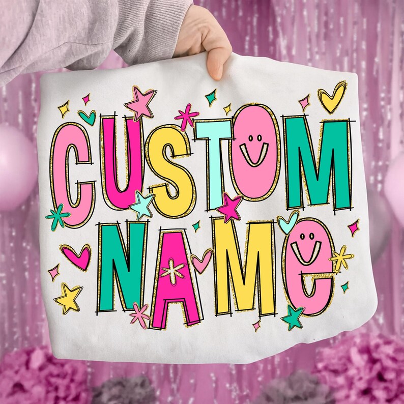 May include: White tote bag with the words "CUSTOM NAME" in colorful, glittery letters. The design includes stars, hearts, and smiley faces. The bag is held up by a person.