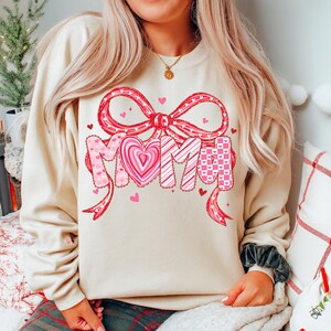 May include: A beige sweatshirt with a large graphic that says "MOM" in pink letters. The "M" letters have heart and bow patterns. A large pink bow is above the letters, and small pink hearts are scattered around the design.