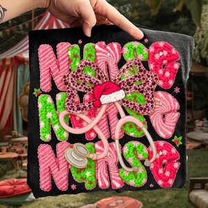 May include: Black t-shirt with the words "NURSE" in pink, green, and white candy cane-style lettering. A bow with a Santa hat and stethoscope are in the center. The bow has a leopard print design. The t-shirt is held up by a hand.