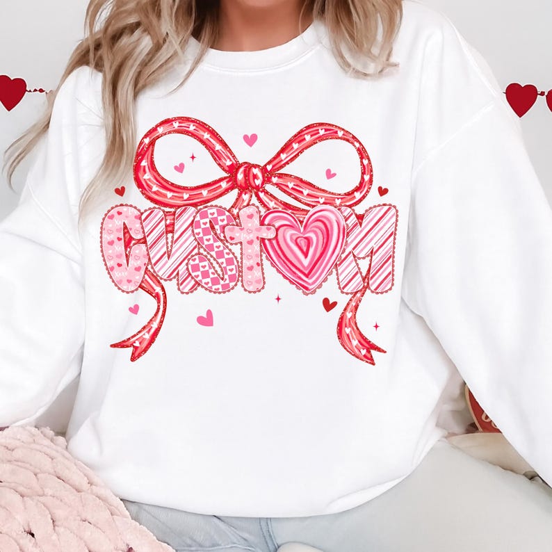 May include: White sweatshirt with a large red bow and the word "CUSTOM" in a heart-themed design. The letters are decorated with pink hearts and stripes. Small pink hearts are scattered around the design, creating a festive look.
