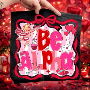 May include: A black tote bag with a Valentine's Day design. The image shows the words "Be Alpha" in red and pink, alongside Cupid, hearts, and a bow and arrow. The bag is held up by a hand.