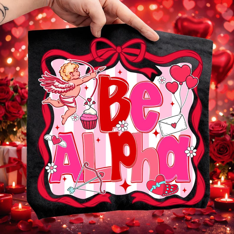 May include: A black tote bag with a Valentine's Day design. The image shows the words "Be Alpha" in red and pink, alongside Cupid, hearts, and a bow and arrow. The bag is held up by a hand.
