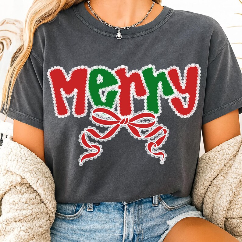 May include: Dark gray t-shirt with the word "MERRY" in red and green, outlined with white lace-like trim. A red bow with white lace detailing is below the word. The shirt is a casual style.