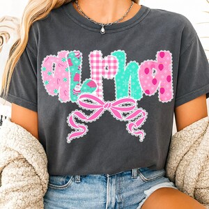Pu&ograve; includere: T-shirt grigio scuro con la scritta "Merry" in un carattere decorativo. Le lettere sono rosa e verde acqua, con un motivo a quadretti e disegni a tema festivo. Un fiocco rosa con un bordo in pizzo &egrave; sotto la scritta.