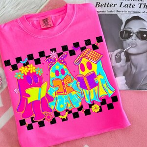 May include: A bright pink t-shirt featuring a colorful graphic of three ghost-like figures reading books. The figures are outlined in black and have yellow, blue, and orange accents. The shirt has a black and white checkered pattern.