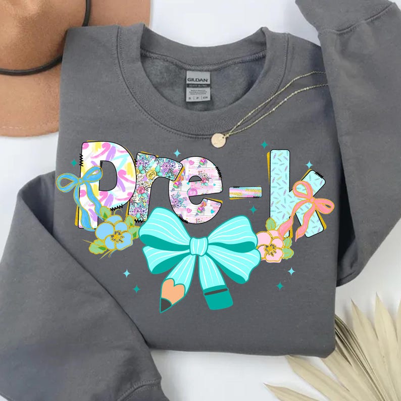 May include: A gray sweatshirt with the words "Pre-K" in a colorful floral design. The letters are decorated with bows, flowers, and a pencil. A gold necklace is draped across the sweatshirt.