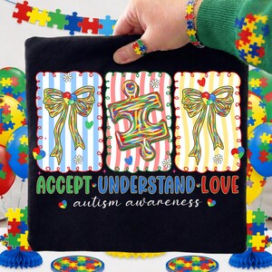 May include: Black t-shirt with three colourful framed designs. The designs include a bow, a puzzle piece, and another bow. The shirt has the words "ACCEPT UNDERSTAND LOVE" and "autism awareness". A hand is holding the shirt.