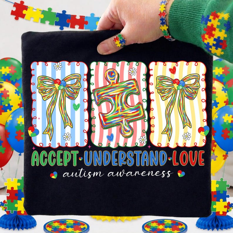 May include: Black t-shirt with three colourful framed designs. The designs include a bow, a puzzle piece, and another bow. The shirt has the words "ACCEPT UNDERSTAND LOVE" and "autism awareness". A hand is holding the shirt.