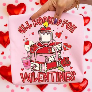 May include: Pink sweatshirt with a Valentine's Day graphic. The design features a stack of books, a candle, a rose, and the text "ALL BOOKED UP FOR VALENTINES". Small hearts and a coffee cup are also included in the design.