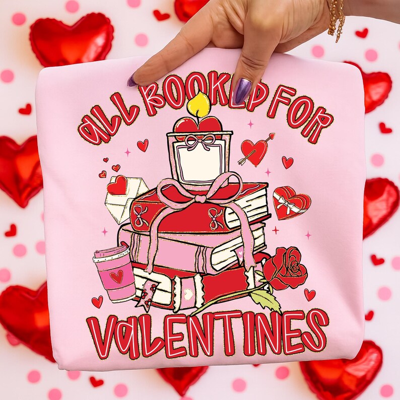 May include: Pink sweatshirt with a Valentine's Day graphic. The design features a stack of books, a candle, a rose, and the text "ALL BOOKED UP FOR VALENTINES". Small hearts and a coffee cup are also included in the design.
