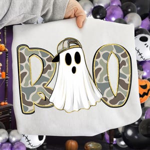 May include: White Halloween-themed sweatshirt with the word "BOO" in a camouflage pattern with gold trim. The "O" features a ghost wearing a camouflage baseball cap. The design is perfect for Halloween.