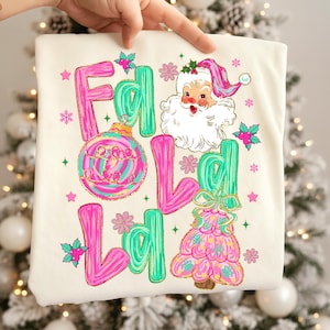 May include: Cream-colored Christmas-themed shirt with the words "Fa La La" in pink and green, along with a Santa Claus graphic and Christmas tree design. The shirt features a festive, colorful design with snowflakes and stars.