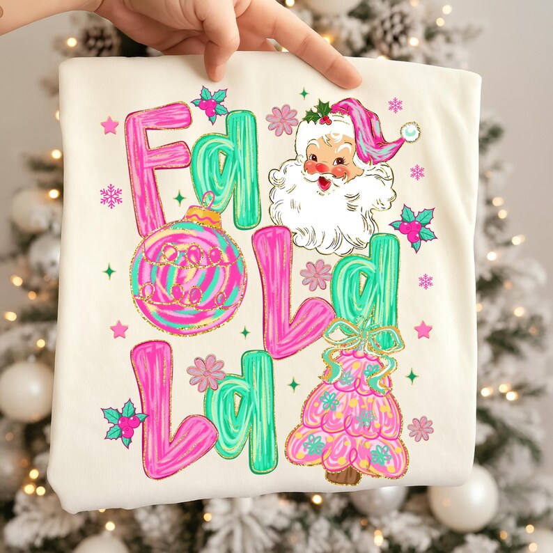 May include: Cream-colored Christmas-themed shirt with the words "Fa La La" in pink and green, along with a Santa Claus graphic and Christmas tree design. The shirt features a festive, colorful design with snowflakes and stars.
