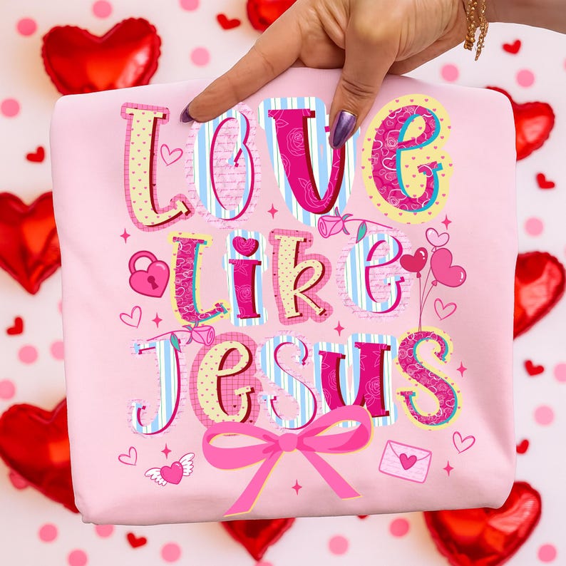 May include: A light pink sweatshirt with the words "LOVE LIKE JESUS" in a colorful, decorative font. The design includes hearts, roses, and a pink bow. The sweatshirt is held up by a hand.