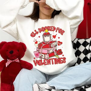 May include: White sweatshirt with a Valentine's Day graphic. The graphic features a stack of books, hearts, and the text "All Booked For Valentines" in red. A red teddy bear is on the left.