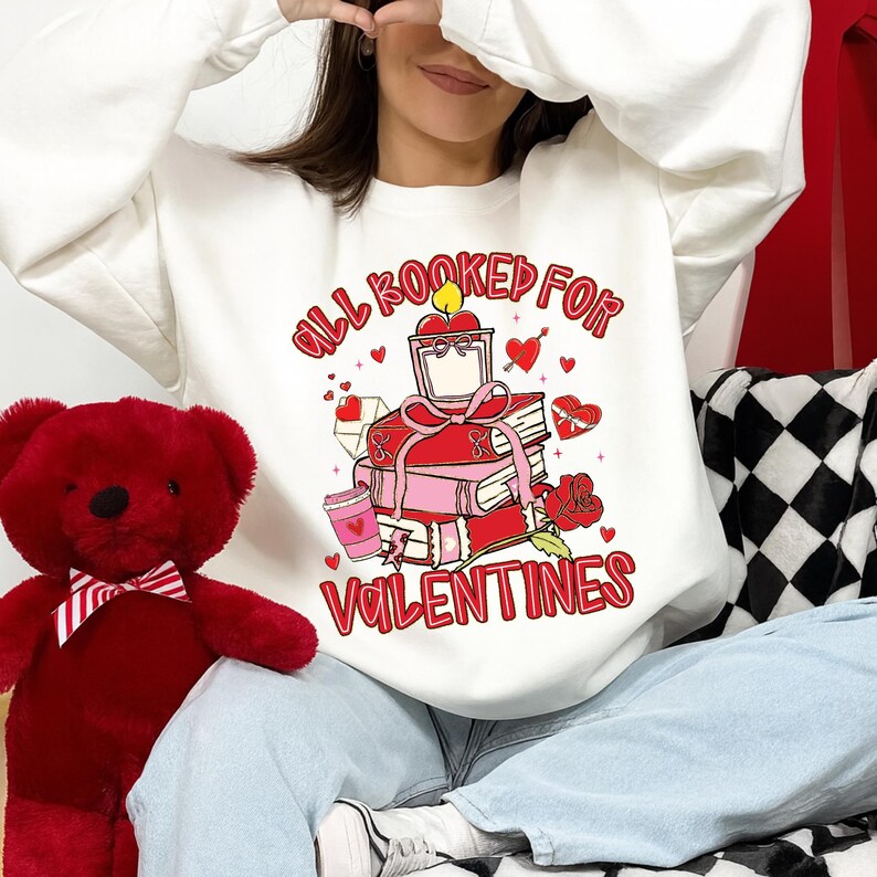 May include: White sweatshirt with a Valentine's Day graphic. The graphic features a stack of books, hearts, and the text "All Booked For Valentines" in red. A red teddy bear is on the left.