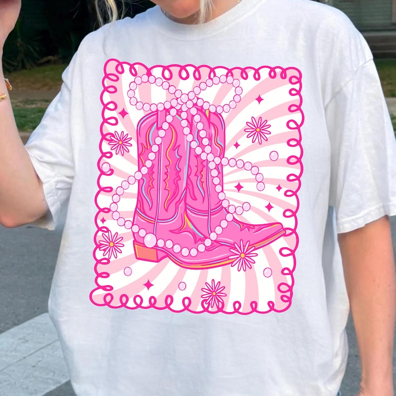 May include: White t-shirt featuring a pink graphic of a cowboy boot adorned with a pearl necklace. The design is set within a pink frame with floral and star accents, creating a playful and feminine aesthetic. The shirt is a casual wear item.