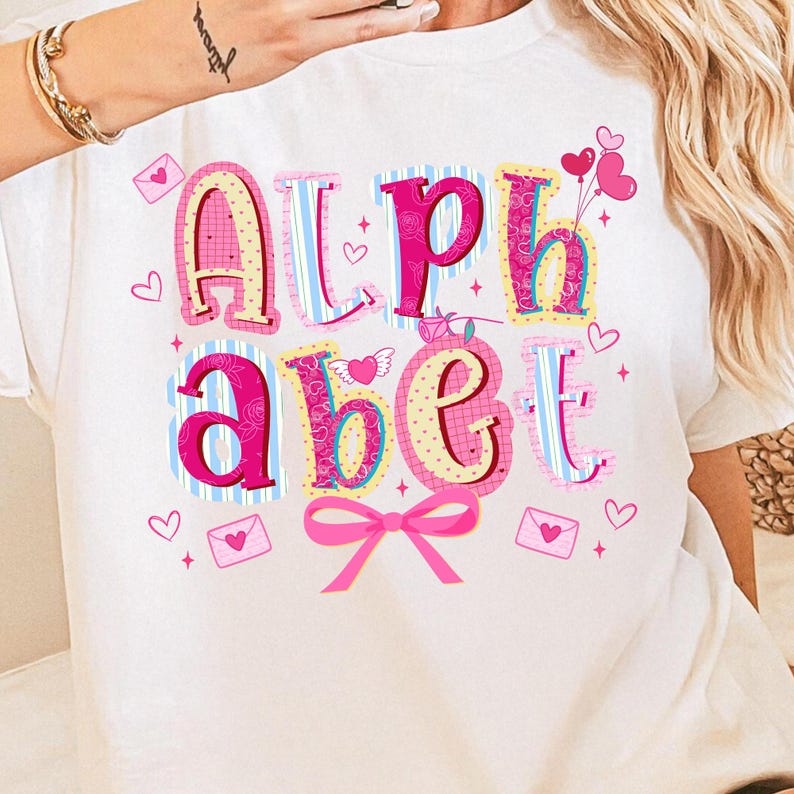 May include: White t-shirt featuring the word "ALPHABET" in a colorful, patchwork design. The letters are pink, blue, and yellow, with hearts, envelopes, and a pink bow. A fun, playful design.
