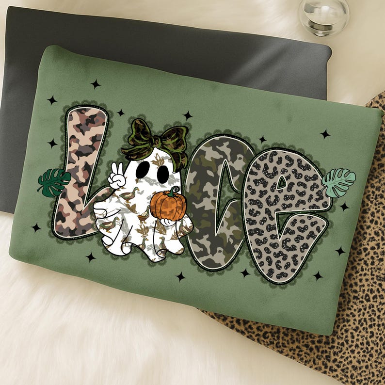 May include: A green pillow with the word "LOVE" in a mix of camouflage and leopard print. A cartoon ghost with a bow and pumpkin is in the center. Black star accents and a lace border complete the design.