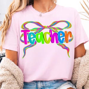 Op de afbeelding: Lichtroze t-shirt met het woord "Teacher" in kleurrijke letters en een regenboog strik ontwerp. Het shirt is gemaakt van zacht materiaal en geschikt voor dagelijks gebruik.