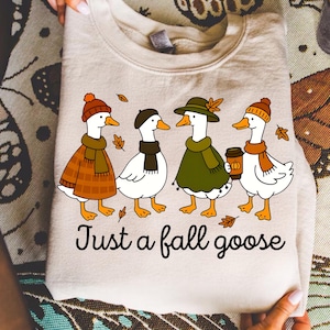 May include: Beige sweatshirt featuring a whimsical illustration of four geese dressed in fall attire, including hats, scarves, and a coffee cup. The text "Just a fall goose" is printed below the geese. A cozy autumn-themed apparel item.