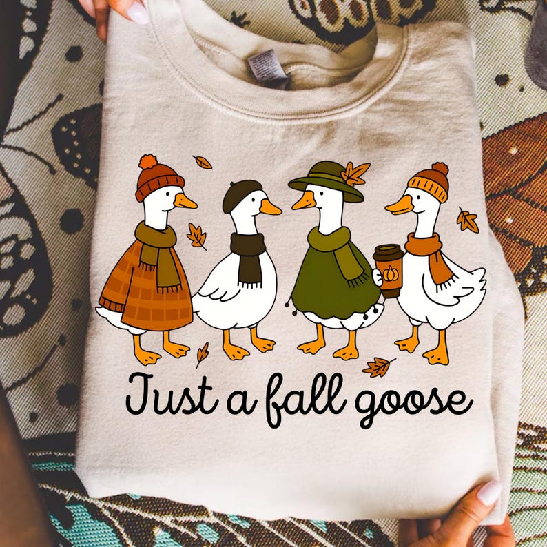 May include: Beige sweatshirt featuring a whimsical illustration of four geese dressed in fall attire, including hats, scarves, and a coffee cup. The text "Just a fall goose" is printed below the geese. A cozy autumn-themed apparel item.