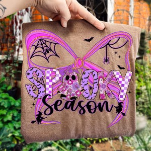 May include: A brown fabric item featuring the words "Spooky Season" in purple and black script. The design includes a purple bow, a floral ghost, bats, spiders, and witch silhouettes, all in a Halloween theme.