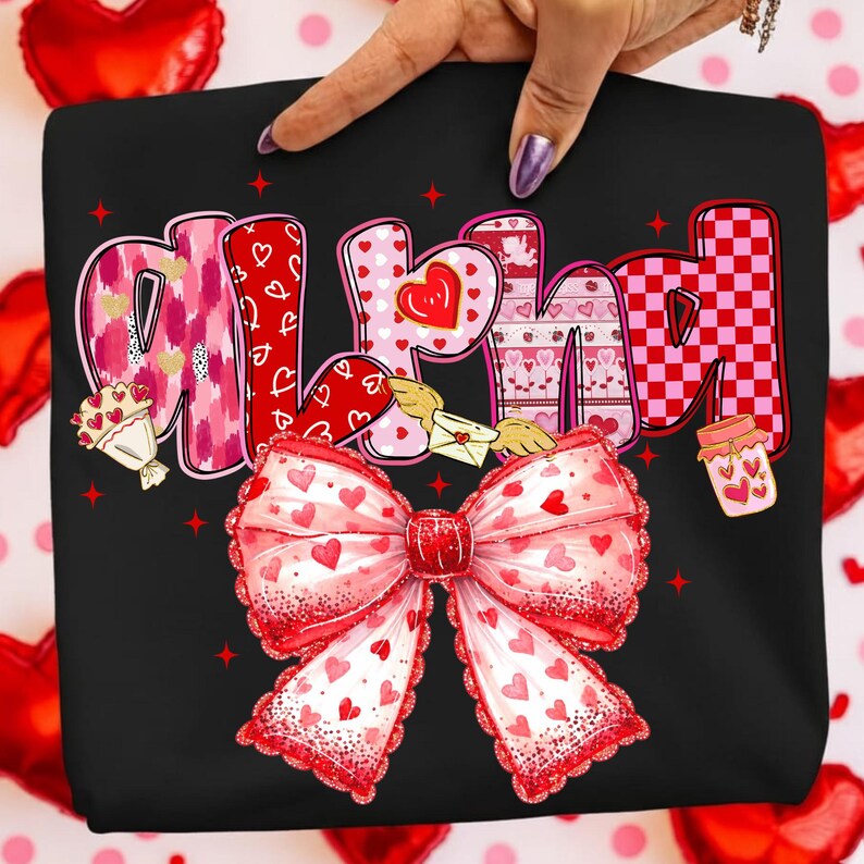 May include: Black sweatshirt with a large, decorative bow at the bottom. The word "MOM" is printed across the top in pink, red, and white letters with heart and Valentine's Day themed designs. A hand is pointing at the sweatshirt.