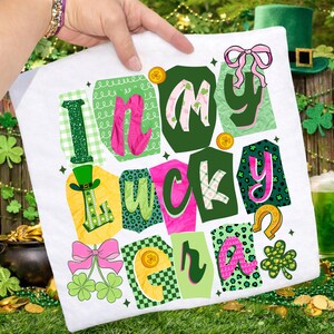 May include: White t-shirt with a colorful graphic design that says "In My Lucky Era." The design features green, pink, and yellow elements, including shamrocks, a horseshoe, and a leprechaun hat. The shirt is held up by a person.