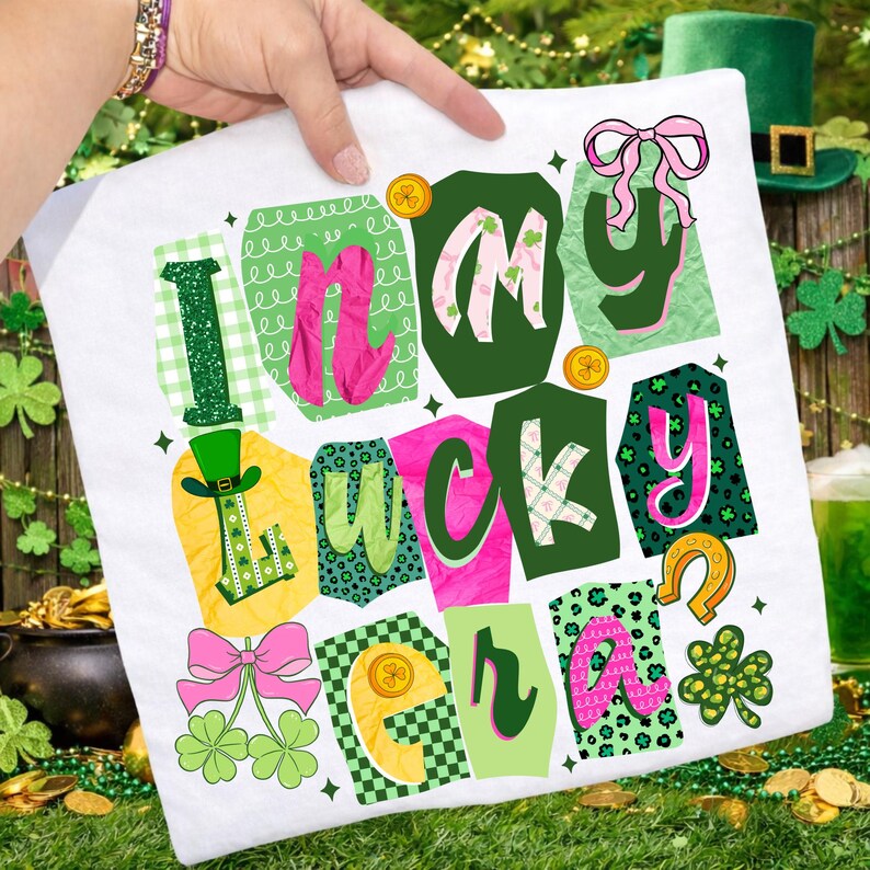 May include: White t-shirt with a colorful graphic design that says "In My Lucky Era." The design features green, pink, and yellow elements, including shamrocks, a horseshoe, and a leprechaun hat. The shirt is held up by a person.