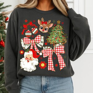 May include: Dark grey sweatshirt with a Christmas-themed design. The graphic features a reindeer, Santa Claus, Christmas tree, baubles, and red and white gingham bows. Red stars and flowers complete the festive look.