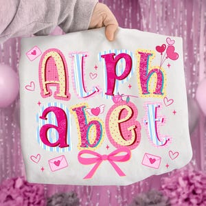 May include: A white tote bag with the word "Alphabet" in pink, yellow, and blue letters. The letters are decorated with hearts, stripes, and polka dots. A pink bow and heart-shaped balloons are also featured.