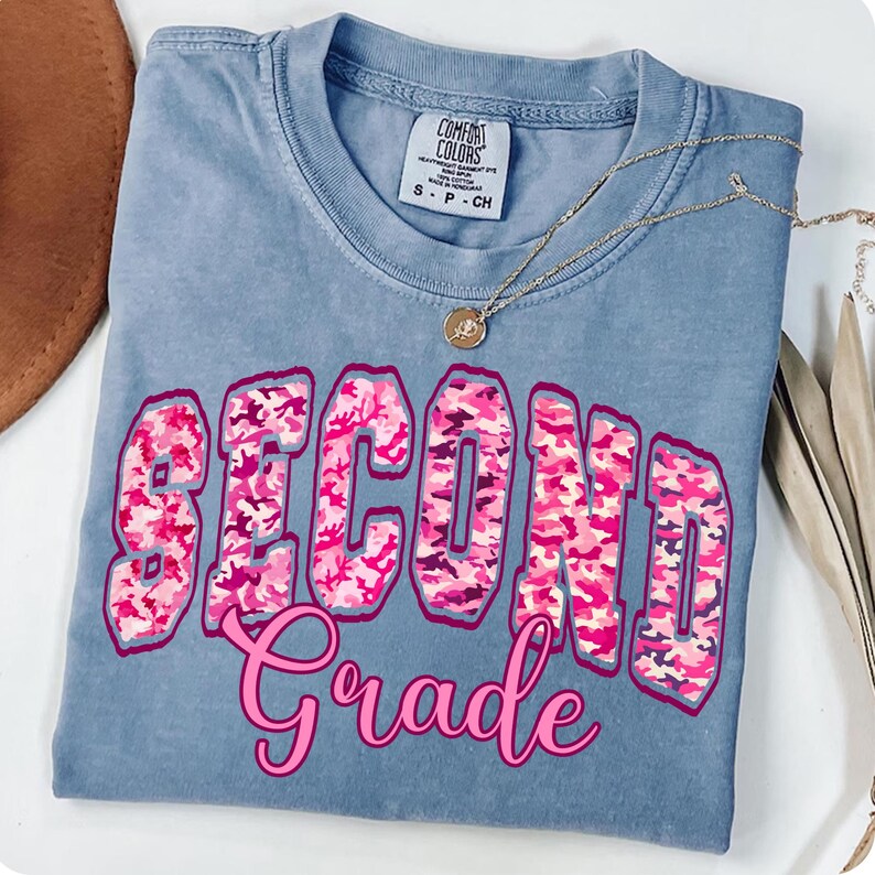 May include: A blue Comfort Colors t-shirt with the words "SECOND Grade" in pink and camouflage print. A gold necklace with a pendant is draped across the shirt. The shirt is folded neatly.