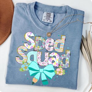 May include: A blue Comfort Colors t-shirt with the words "Sped Squad" in colorful, patterned letters. The design includes a turquoise bow, a pencil, and floral accents. A gold necklace rests on the shirt.