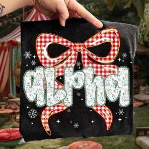 May include: Black t-shirt featuring a large red and white gingham bow design with gold trim. The word "Alpha" is printed in a floral font. Snowflakes and stars are scattered around the design.
