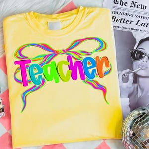 Op de afbeelding: Geel T-shirt met het woord "Teacher" in kleurrijke letters, geaccentueerd door een regenboogstrikontwerp. Het shirt is opgevouwen en gepresenteerd op een roze en wit gestreept oppervlak.