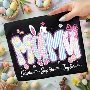 May include: Black t-shirt with the word "MOM" in a patchwork design, bunny ears, and the names Olivia, Sophia, and Taylor. The shirt has a decorative lace trim and Easter-themed accents, including eggs and butterflies.