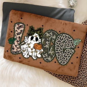 May include: A brown decorative pillow with the word "LOVE" in a whimsical design. The "L" and "E" have a camouflage pattern, while the "O" has a leopard print. A ghost with a bow and pumpkin is in the center. Black star accents are scattered around the letters.