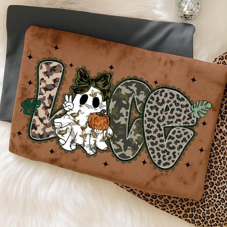 May include: A brown decorative pillow with the word "LOVE" in a whimsical design. The "L" and "E" have a camouflage pattern, while the "O" has a leopard print. A ghost with a bow and pumpkin is in the center. Black star accents are scattered around the letters.