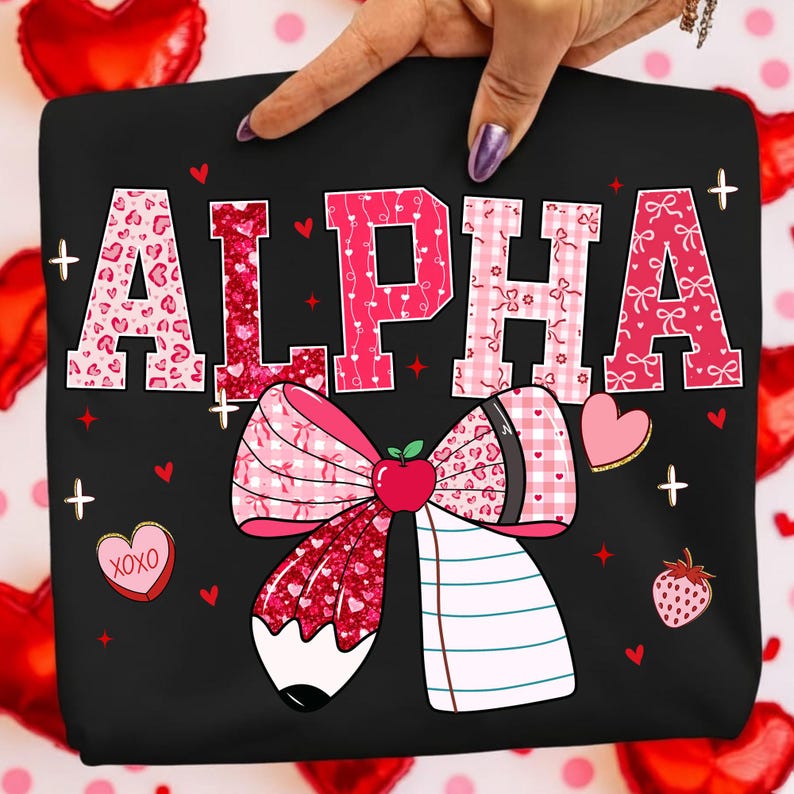 Op de afbeelding: Zwart t-shirt met het woord "ALPHA" in roze, versierd met hart- en strikpatronen. Een grote strik met een potlood en notitieboek staat eronder. Kleine hartjes, aardbeien en de tekst "XOXO" zijn ook aanwezig.