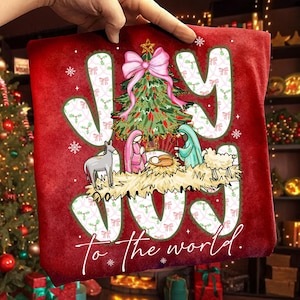 May include: Red Christmas-themed pillow with the words "JOY" and "to the world" in white. The design includes a Christmas tree, nativity scene, and snowflakes. A hand is holding the pillow.