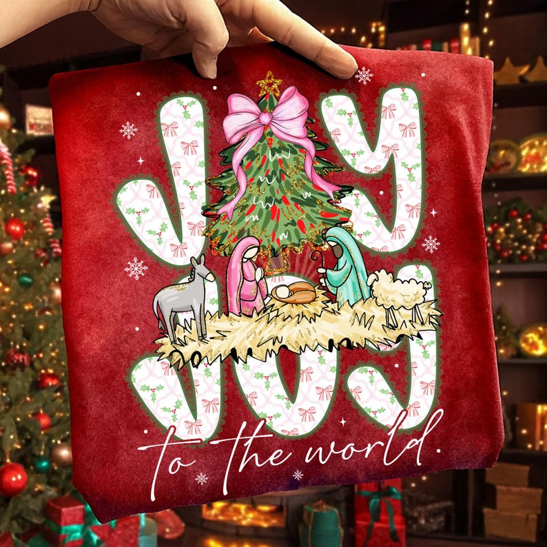 May include: Red Christmas-themed pillow with the words "JOY" and "to the world" in white. The design includes a Christmas tree, nativity scene, and snowflakes. A hand is holding the pillow.