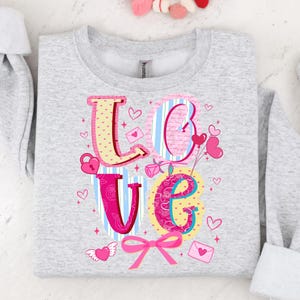 以下が含まれることがあります： カラフルなデザインで「LOVE」の文字がプリントされたライトグレーのスウェットシャツ。文字はハート、ストライプ、花柄で装飾されています。ピンクのリボンとハート型の風船がバレンタインデーのテーマを完成させています。
