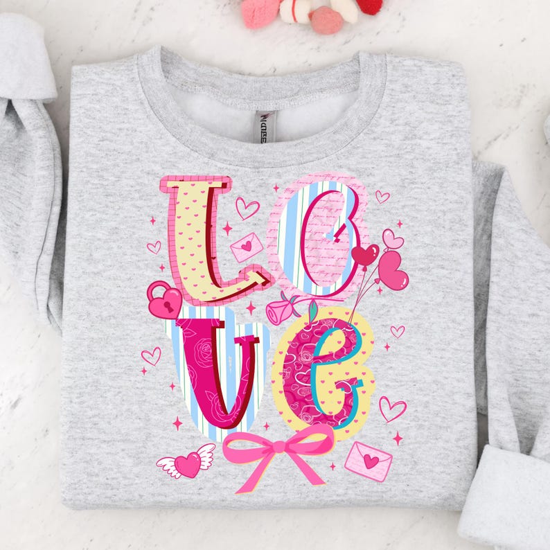 以下が含まれることがあります： カラフルなデザインで「LOVE」の文字がプリントされたライトグレーのスウェットシャツ。文字はハート、ストライプ、花柄で装飾されています。ピンクのリボンとハート型の風船がバレンタインデーのテーマを完成させています。