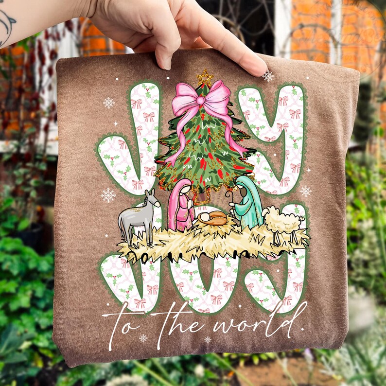 May include: A brown Christmas-themed decorative pillow with the words "Joy to the world." The design includes a Christmas tree with a pink bow, the nativity scene, and the word "JOY" in a decorative font.