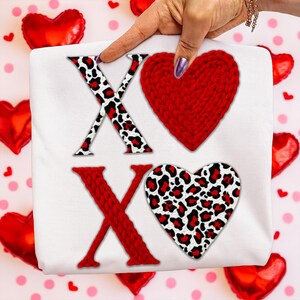 Może przedstawiać: Biała tkanina z literami "XO". Litera "X" jest czerwona ze wzorem w panterkę, a "O" to czerwone dziergane serce. Drugie "X" jest czerwone, a drugie "O" to serce w panterkę. Czerwone balony w kształcie serca w tle.