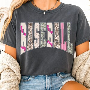 May include: Dark gray t-shirt with the word "BASEBALL" in a bold, decorative font. The letters feature baseball-themed patterns, including pink stitching, leopard print, and camouflage. The shirt has a crew neck and short sleeves.